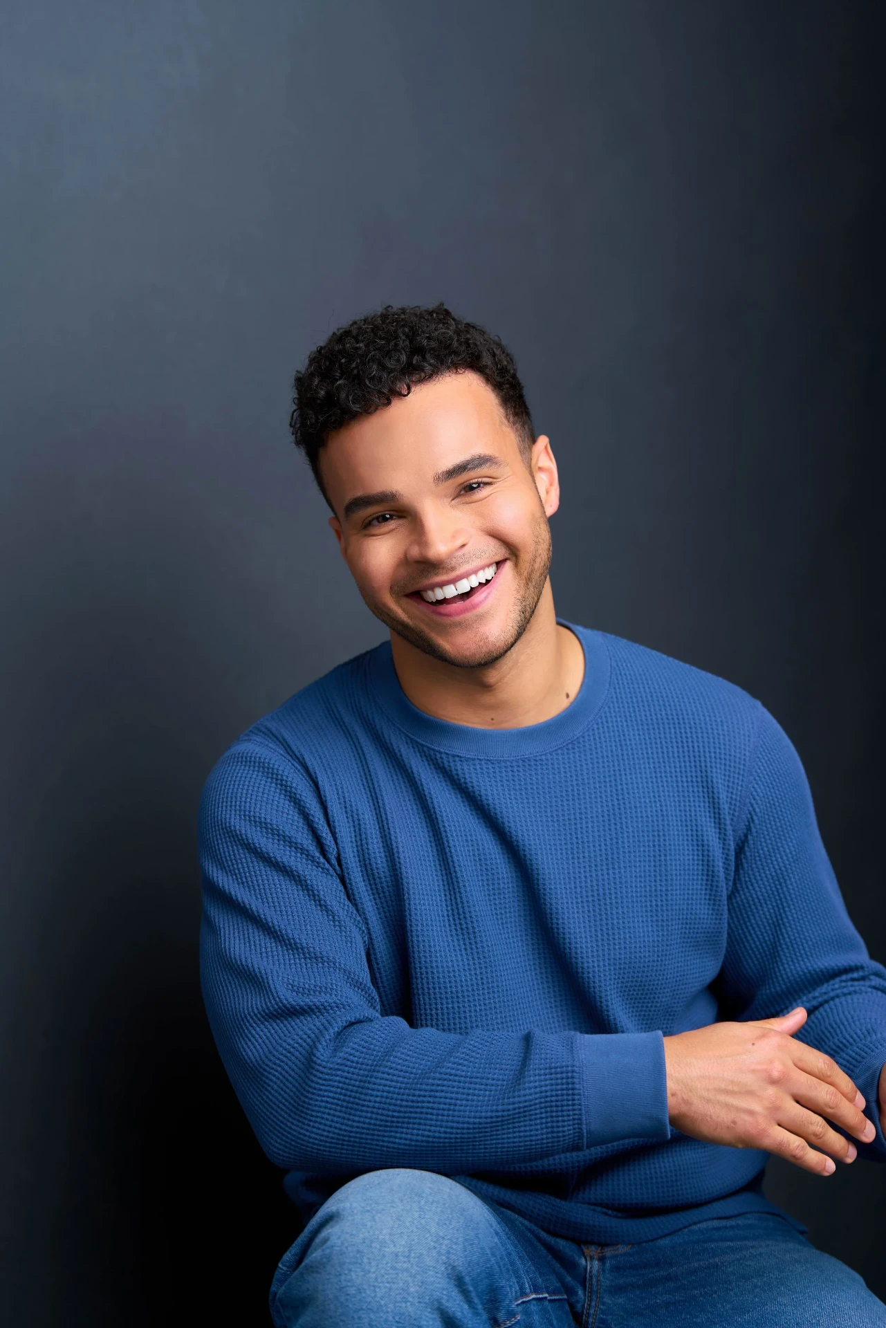 Editorial actor headshot of smiling actor in a blue sweater seated on a dark backdrop, photographed by an Atlanta actor headshot photographer